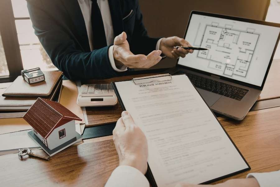 Un conseiller immobilier présente les plans d'un immeuble de rapport à un investisseur autour d'un contrat d'achat et d'une maquette de maison.