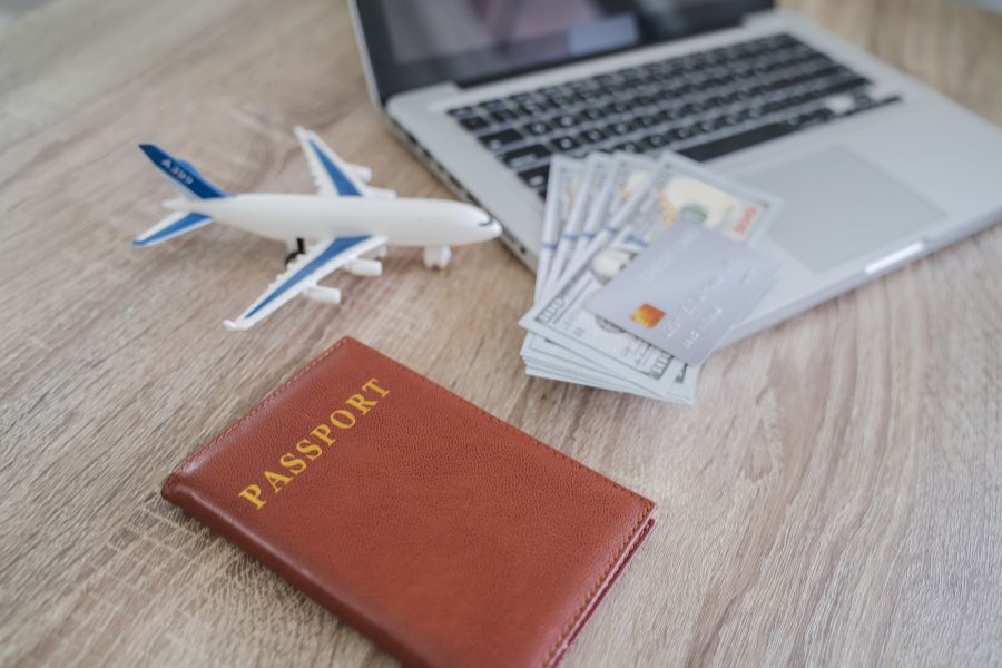 Un passeport marron, un avion miniature, des dollars et une carte bancaire posés sur un ordinateur portable, évoquant la préparation d'un voyage longue durée avec assurance.